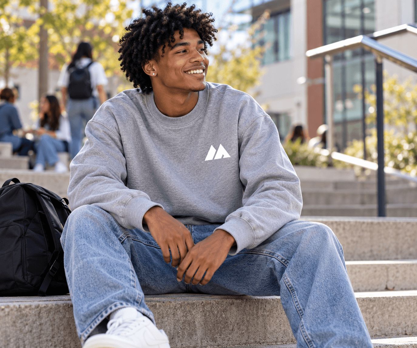 Smiling student in a grey MAIS Apparel crewneck sweatshirt sitting on campus steps with a backpack